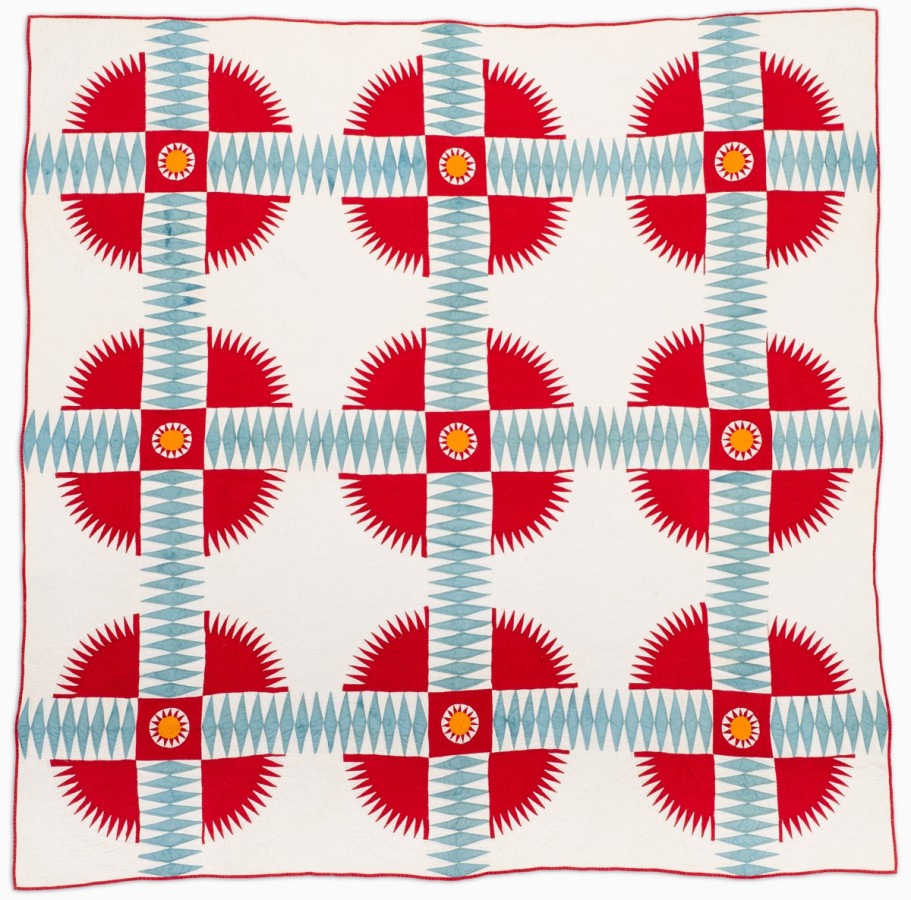 This quilt is symmetrical with a white background and red trim. There are three designs vertically and three horizontally. The design is the same in every section. In the center of the design is a yellow sun with red sun rays. Behind the sun is a white circle inside a red square. Around the square are four large, red triangles with a saw tooth edge. These designs are separated, but create a circle. Separating the saw tooth pieces are horizontal and vertical lines of light blue think diamonds.