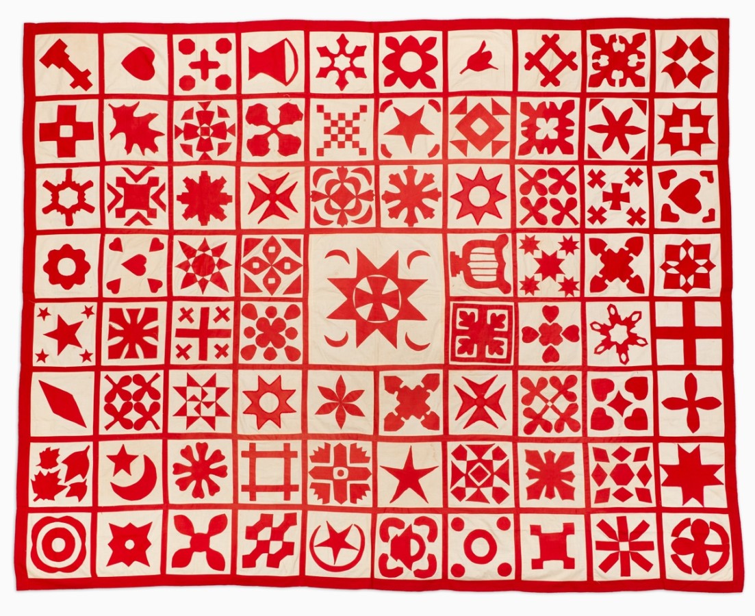 This American quilt's color scheme is red and white. All the squares have a red outline and design in the center and the background is white. Vertically there are eight squares and horizontally there are ten squares. In the center there is one large square (the size of four square combined). Each square has a different design.